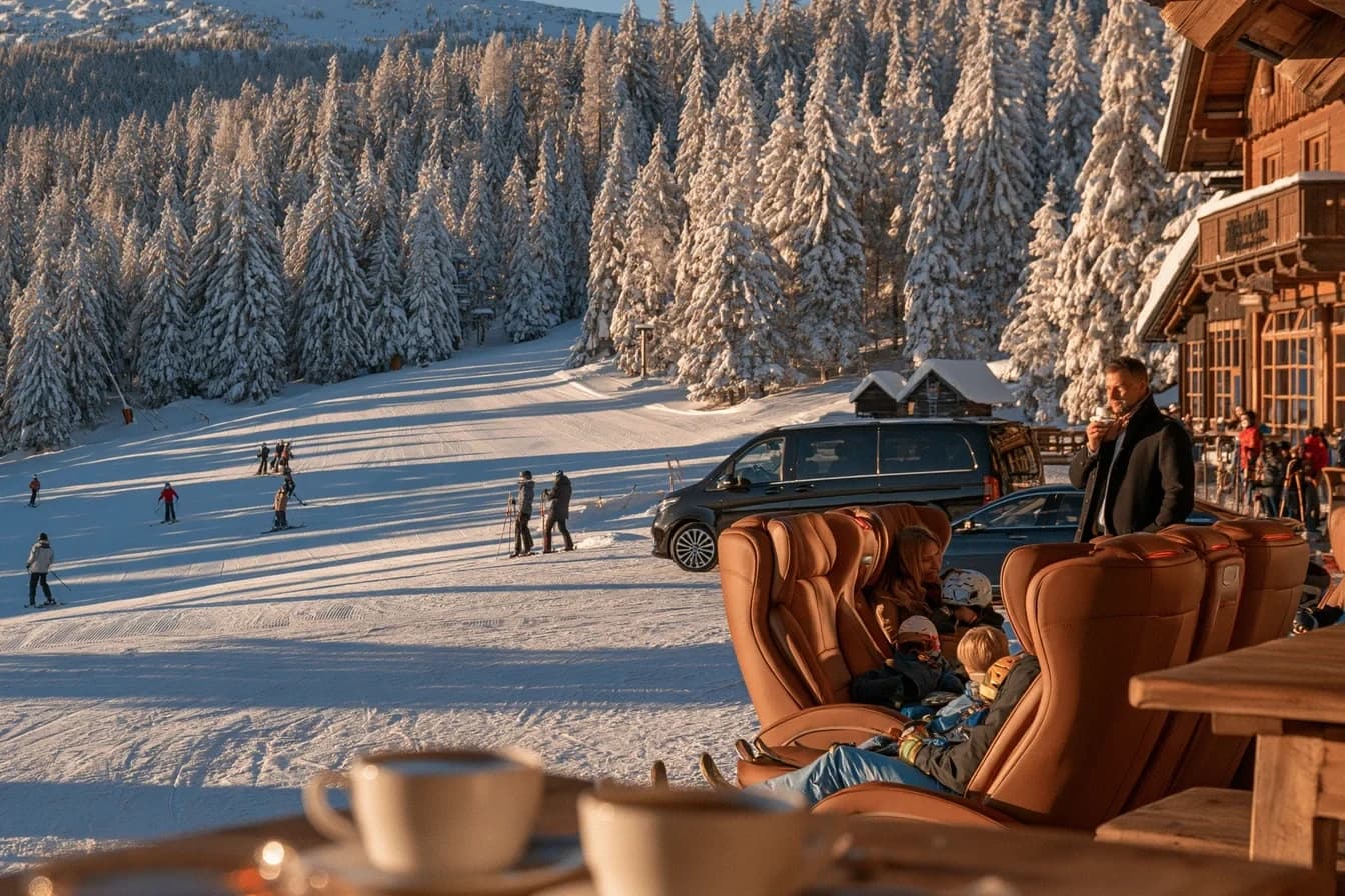 Sunny ski slope with snowy spruce forest seen from a wooden alpine hut terrace, with a black premium van and chauffeur waiting nearby